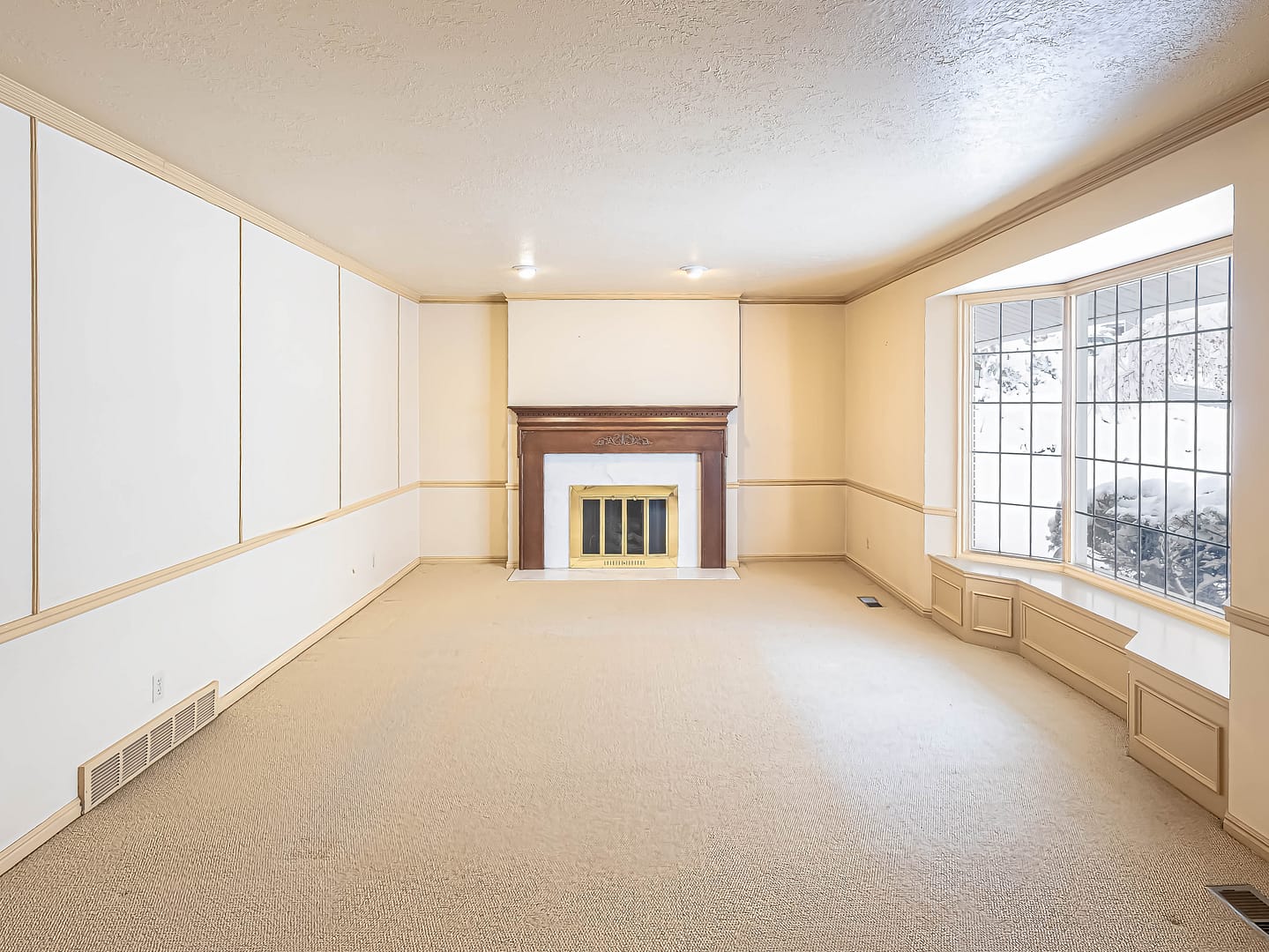 A vacant living room featuring a beige carpet, a fireplace with a wooden mantle, and a large bay window with grid panes.